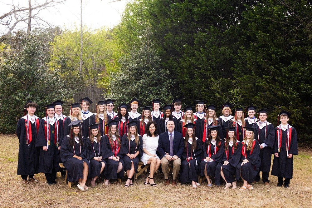 Heart transplant recipient John A Lee is pictured pre-transplant in an outdoor graduation photo for Konos Academy where he is principal and CEO. He is seated in the front row with his wife. There are about 30 young adults in graduation robes and caps. John has light skin, short dark hair, and a gray beard and he wears a suit. His wife has light skin, red lipstick, curly shoulder-length dark hair, and a white knee-length dress.