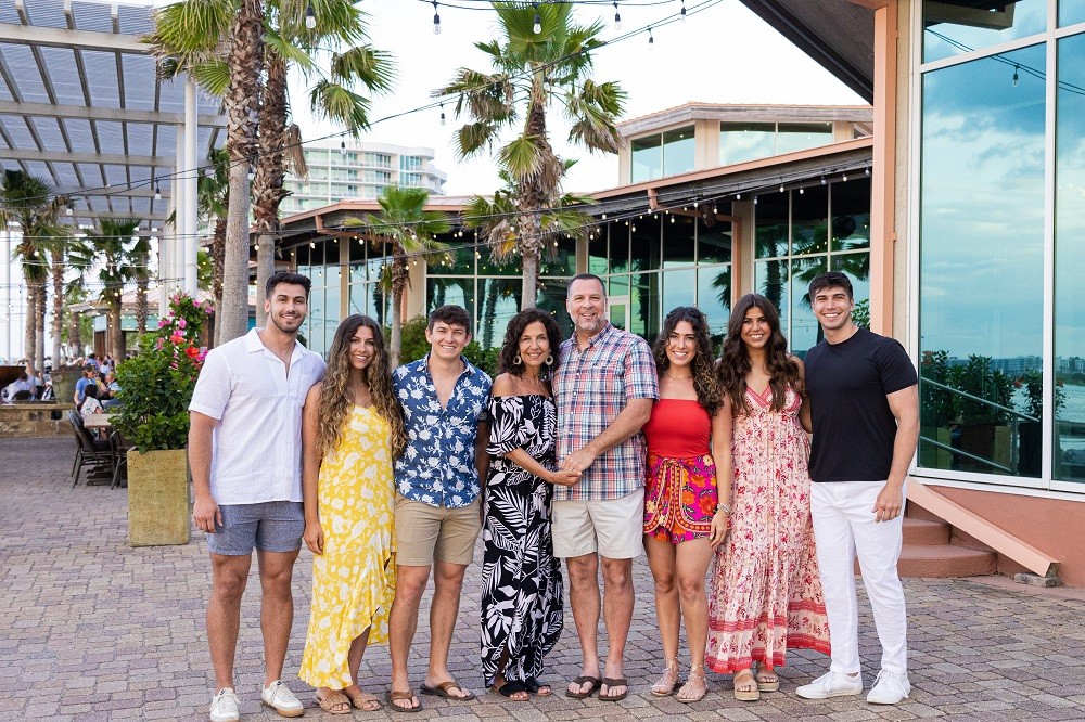 Heart transplant recipient John A Lee is pictured pre-transplant with his wife and three women and three men, including his five children. They are outside in a courtyard with palm trees and outdoor seating. All are dressed in summery clothing such as flowing floral dresses. John has light skin, short dark hair, and a short gray beard and he wears khaki shorts and sandals with a short-sleeved plaid button-up shirt.