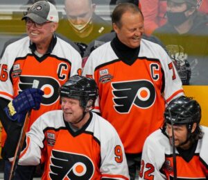 A photo of four Flyers Alumni Association members smiling on the ice in their hockey gear with the black, white, and bright orange Philadelphia Flyers logo and branding.