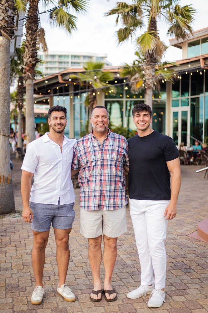 Heart transplant recipient John A Lee is pictured pre-transplant in an outdoor courtyard setting with his two sons. John has light skin, short dark hair, and a short gray beard and we wears khaki shorts, sandals, and a plaid short sleeved button up shirt. Both of his sons have short styled dark hair, light skin, and dark eyes.