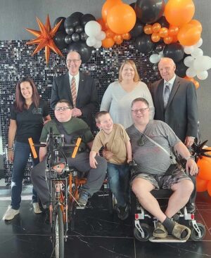 Dylan McDonnell and his family with Help Hope Live Executive Director Kelly L Green as Dylan sits on his new Flyers themed orange adaptive bike. Dylan is a teen with light skin and glasses and short brown hair. Kelly L Green has light skin, red-brown hair past her shoulders, and a black Help Hope Live polo. With them are Brad Marsh and Bob Kelly, Flyers Alumni members.