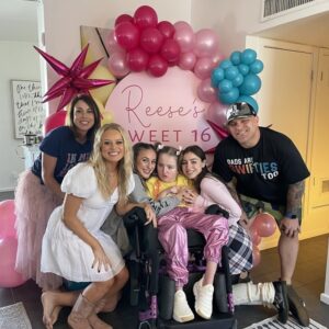 At her sweet 16 party, Reese is 16-year-old Reese D'Ortenzio is surrounded by four woman including her mom and 1 man, her dad, with lots of pink, blue, and yellow birthday balloons. She has light skin, brown hair in 2 pigtails, pink sparkling pants, and white sneakers. She is seated in her black mobility device. Her mom has light skin, shoulder-length straight brown hair, a pink flowing skirt, and a shirt that reads In My Lover Era in groovy letters. Her dad has light skin, glitter on his cheeks, a ball cap, camo shorts, and a shirt that reads Dads Are Swifties Too.