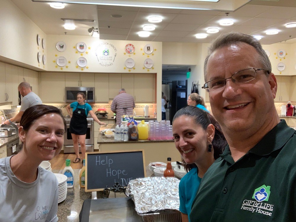 Heart transplant recipient Bill Soloway joins Help Hope Live team members Shannon and Kelly L Green at the Gift of Life Howie's House Home Cook Heroes kitchen. Bill has light skin, short gray hair, a Gift of Life polo, and glasses. Shannon has light skin and brown hair. Kelly L Green has light skin and black hair. They are in a kitchen with a chalkboard sign that reads Help Hope Live.