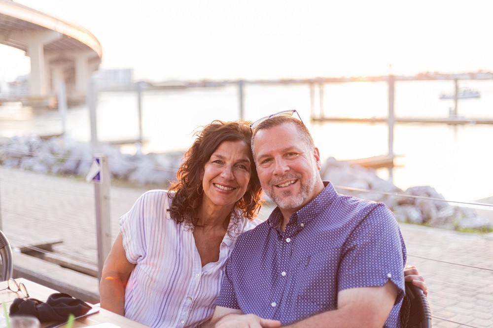 In a photo at an outdoor restaurant seating area that is suffused with sunset golden light, heart transplant recipient John A Lee is pictured pre-transplant with his wife. John has short gray hair, a short gray beard, dark eyes, light skin, and a blue patterned short sleeved button up shirt. His wife has light skin, dark eyes, gold hoop earrings, curly brown hair to her shoulders, and a breezy pastel striped white short sleeved button up shirt.