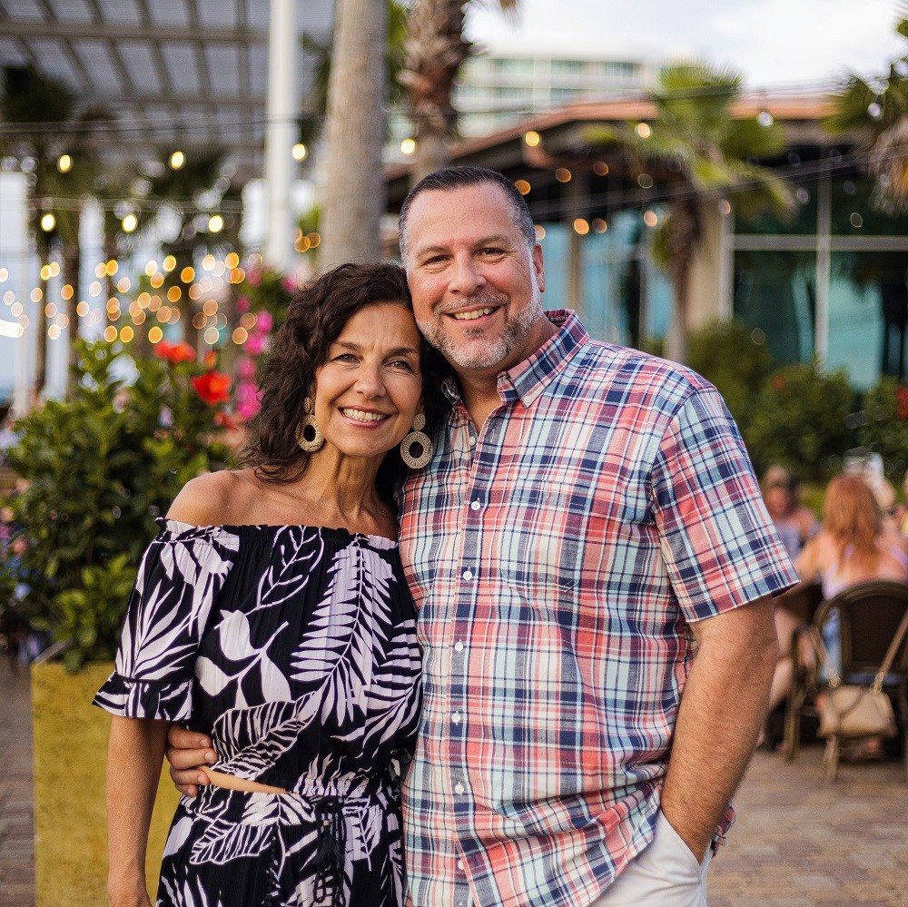 In an outdoor courtyard setting, John A Lee is pictured before his heart transplant with his wife. He has light skin, short dark hair, and a short gray beard and he wears khaki shorts and a plaid button-up short sleeved shirt. His wife has light skin, curly brown hair to her shoulders, hazel eyes, decorative circular gold earrings, and a two-piece summery outfit with white plant silhouettes on black breezy fabric.