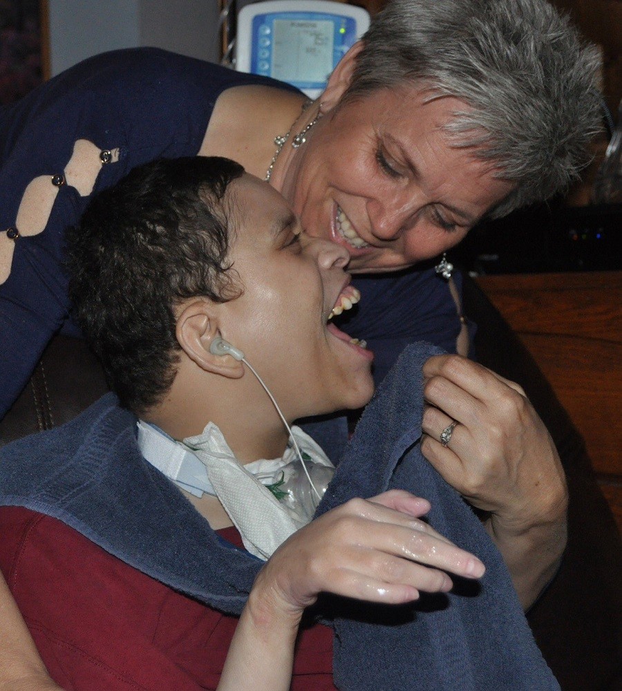 Donivan Hicks leans over to grin at his mom, Angie, who smiles back at him. Donivan is seated in a wheelchair and has a breathing tube in his neck. He has light brown skin and short dark hair with a navy towel around his neck that Angie is raising to his face. Angie has light skin, short gray hair, and a blue long-sleeved shirt.