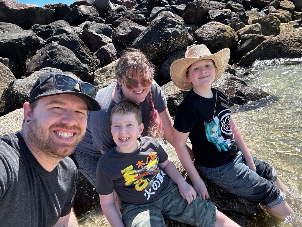 A family photo with Carson, his older brother, his mom, and his dad seated near an outcropping of rocks in a foot of crystal clear water leading out to a bay or ocean. They are all dressed comfortably in gray or black t-shirts and rolled-up pants. Dad has light skin, black gauges in his ears, a ball cap, and a short brown beard. Mom has light skin, aviator sunglasses, and braided partially pink-dyed brown hair with bangs. Carson light skin skin and short brown hair. His older brother has light skin and a straw sunhat.