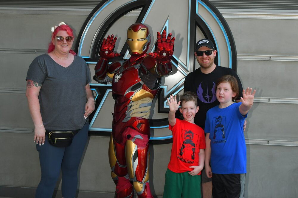 Carson Tussing, his older brother, and his mom and dad pose next to someone dressed in a very elaborate Ironman superhero costume in metallic red, gold, and silver. Carson and his brother both extend a hand to replicate an Ironman hero pose. Carson's mother has light skin, bright pink hair, and sunglasses. Carson has light skin, short brown hair, and a red Mandalorian t-shirt. His brother, standing several inches taller, has light skin, longer straight red-brown hair, and a blue Mandalorian t-shirt. Their father behind them has light skin, sunglasses, a ball cap, and a black and purple Star Wars or Mandalorian themed t-shirt.