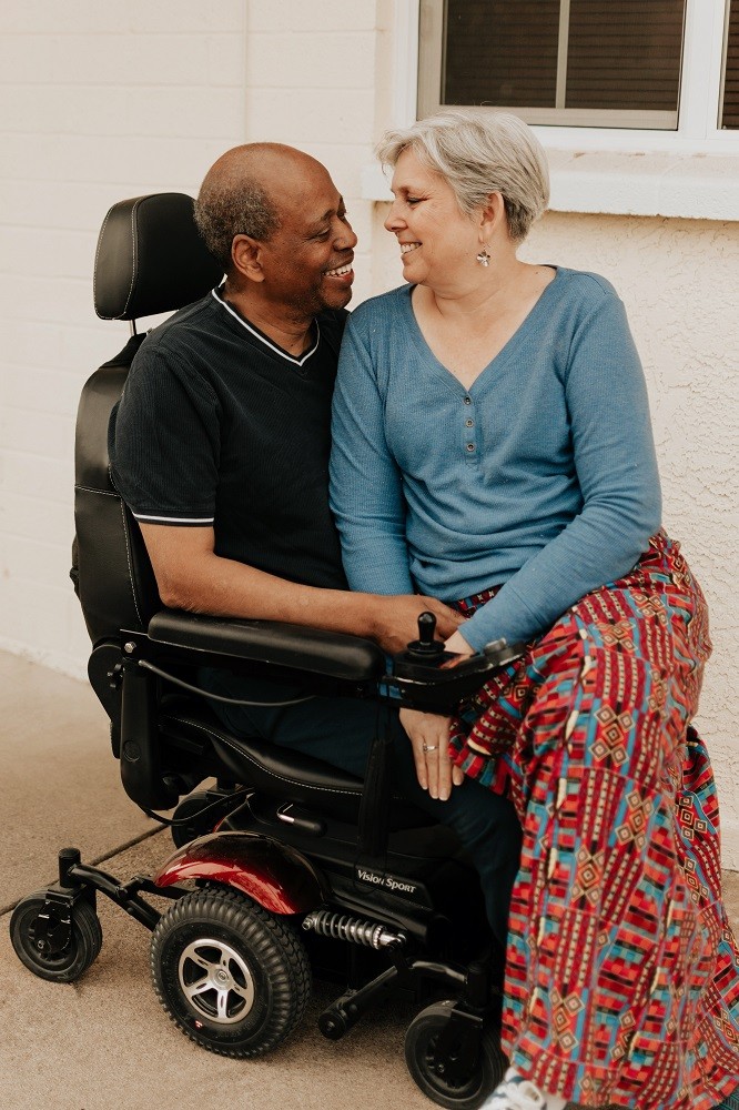 Angie Hicks sits on the lap of her husband Michael Hicks, who is seated in a black power chair. Michael has brown skin and short gray and black hair. Angie has light skin and short gray hair. They are gazing at each other with big smiles.