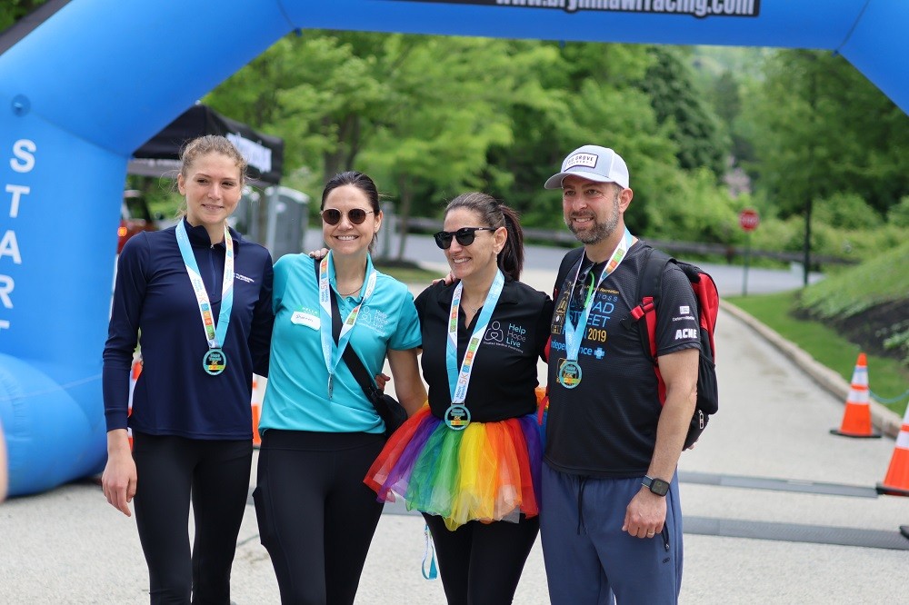 Second place Hope Travels 5K 2024 finisher Cecilia Castelli with two Help Hope Live staff members and a male participant at the finish line outdoors. Cecilia has light skin and curly red-brown hair tied back and wears athletic gear. Director of Communications Shannon has light skin, dark hair pulled back, sunglasses, and a teal Hope Travels t-shirt. Help Hope Live Executive Director Kelly L Green has light skin, dark hair pulled back, sunglasses, a black Help Hope Live polo shirt, and a rainbow tutu. The male participant has light skin, a gray beard, a ball cap, and athletic gear with a Broad Street Run 2019 shirt. All four wear Hope Travels 5K medals.