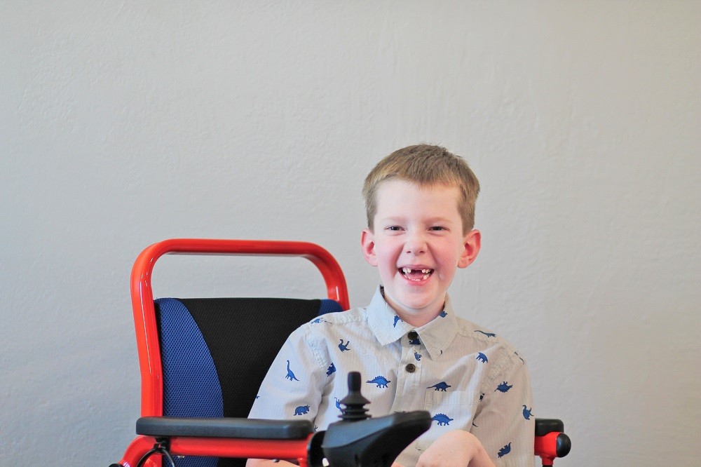 Carson Tussing is seated in a red and blue electric mobility device. He has light skin, his two front teeth missing, and short light brown hair and he wears a collared short-sleeved shirt with dinosaurs printed on it.