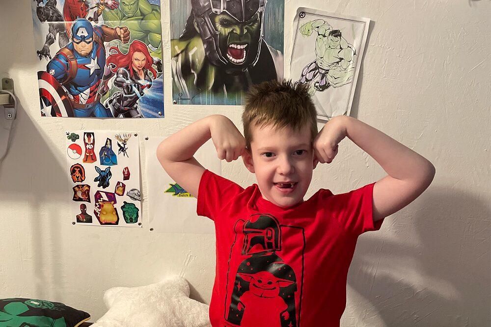 A young Carson Tussing shows off his muscles as he flexes in his bedroom wearing a red Mandalorian-themed t-shirt with anime and superhero posters behind him, especially Hulk. He has light skin, short brown hair, and his two front teeth missing.