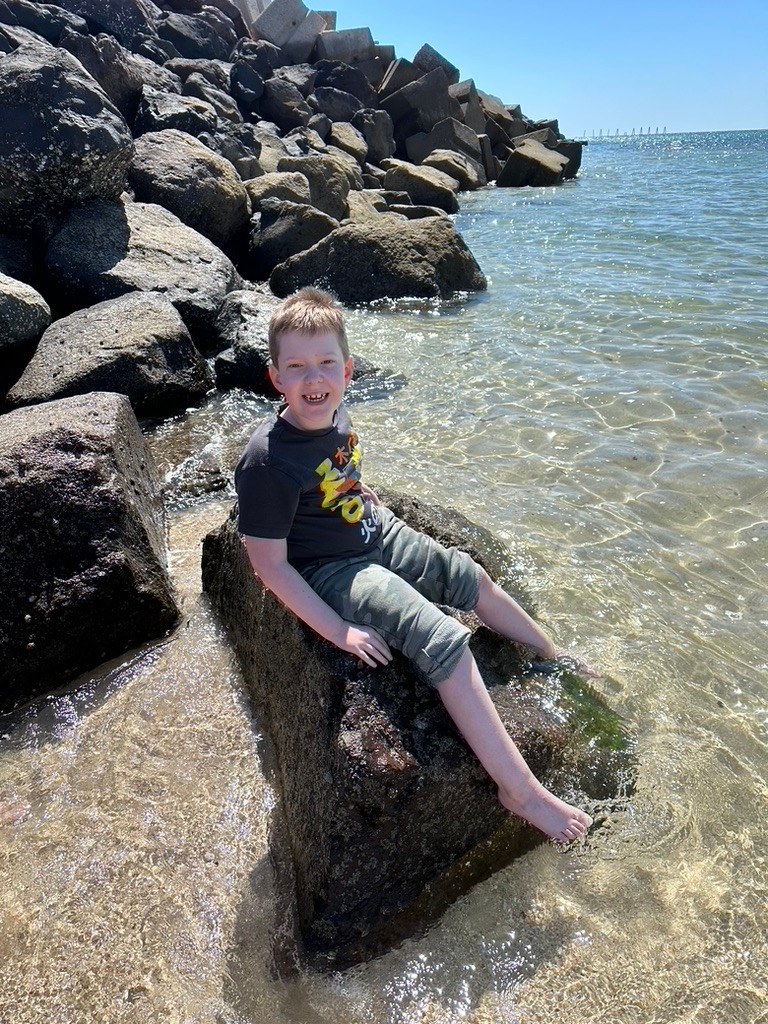 Carson is seated on a rock jutting out of a foot of crystal clear water next to a larger outcropping of rocks leading into an ocean or bay. He has light brown hair, light skin, a black t-shirt with Asian characters on it, and camo printed pants rolled up to his knees.