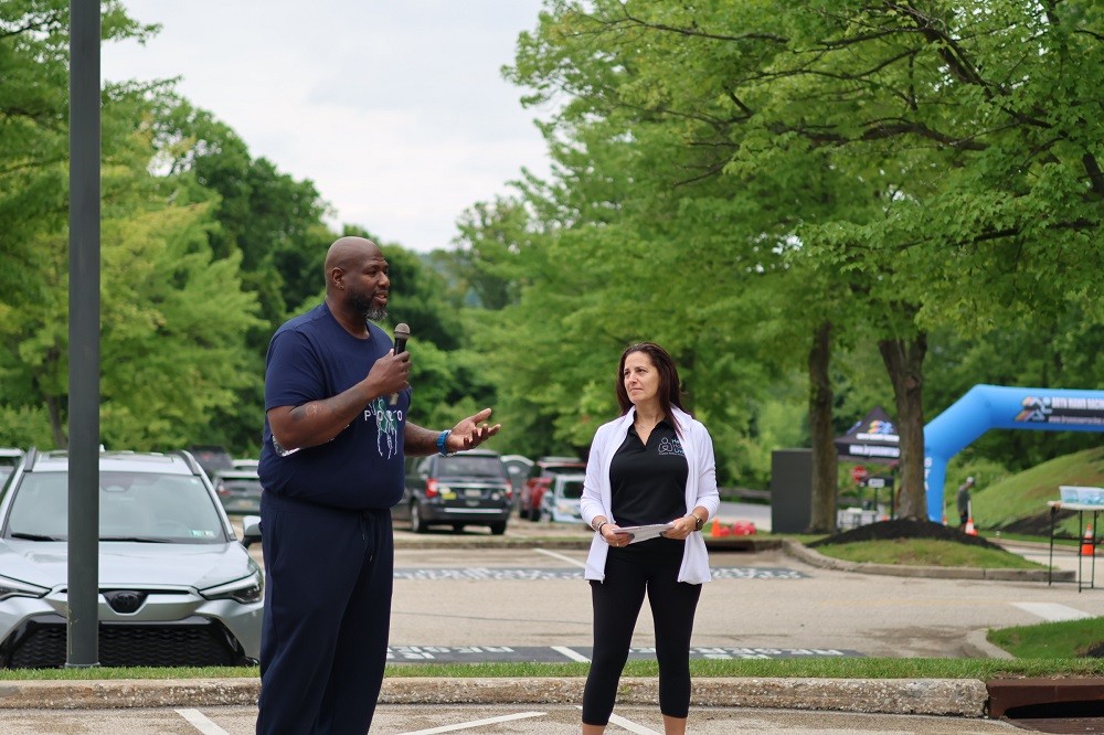 Outdoors at Hope Travels 2024, Help Hope Live Executive Director Kelly L Green looks on as Barrett Brooks speaks with a microphone. Kelly has light skin, dark hair past her shoulders, and a black Help Hope Live polo. Barrett has brown skin and a short black and gray goatee and he stands about 2 feet taller than Kelly.