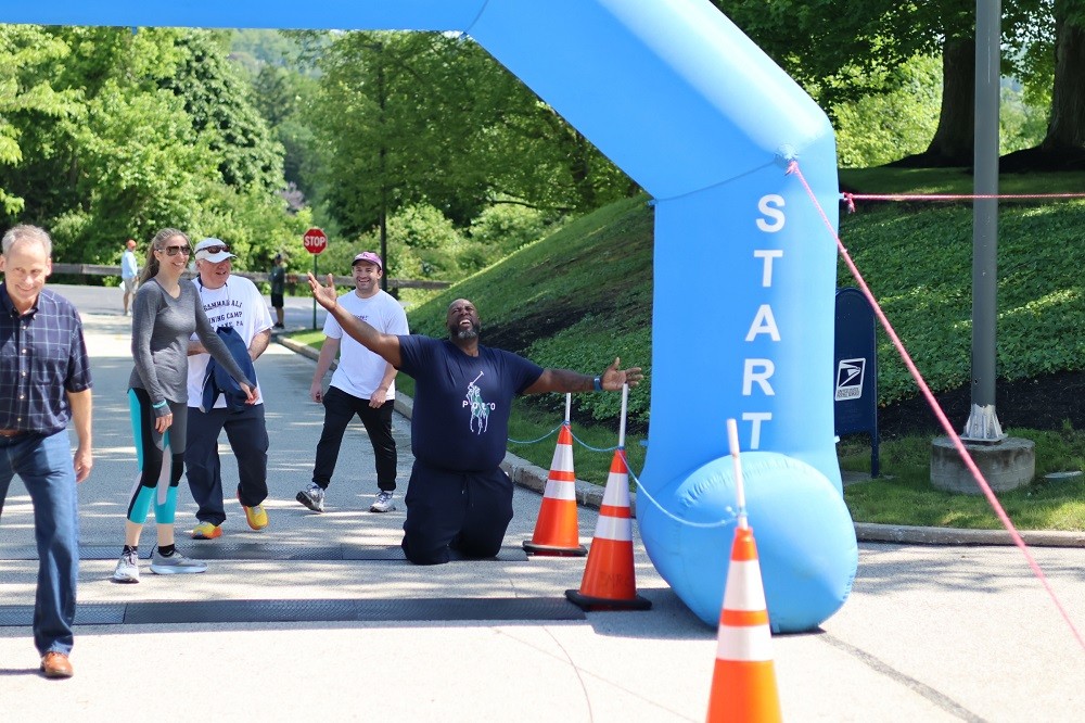 In a funny photo, Barret Brooks is on his knees reaching upward in an expression of relief and endurance as he reaches the finish line of the Hope Travels 5K event in 2024. Surrounded by other participants in athletic gear of various ages who are laughing at his display, Barrett has brown skin and wears a navy t-shirt.