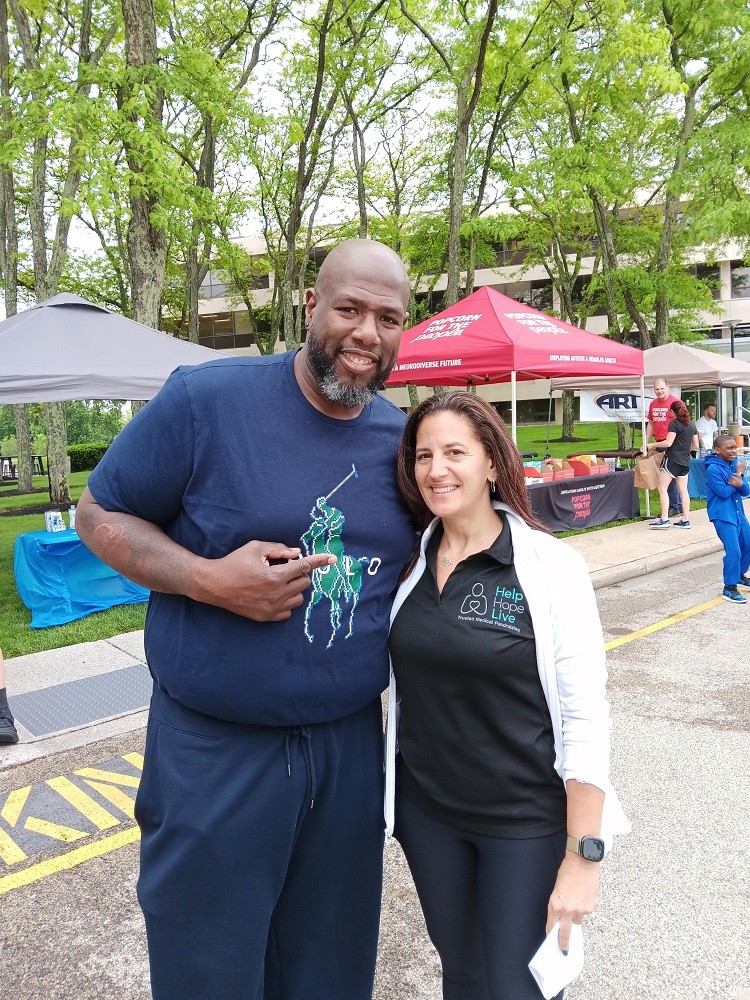 Outdoors at Hope Travels 2024, Help Hope Live Executive Director Kelly L Green poses with Barrett Brooks. Kelly has light skin, dark hair past her shoulders, and a black Help Hope Live polo. Barrett has brown skin and a short black and gray goatee and he stands about 2 feet taller than Kelly in a navy blue t-shirt and navy sweatpants. He is pointing to Kelly with his right index finger.