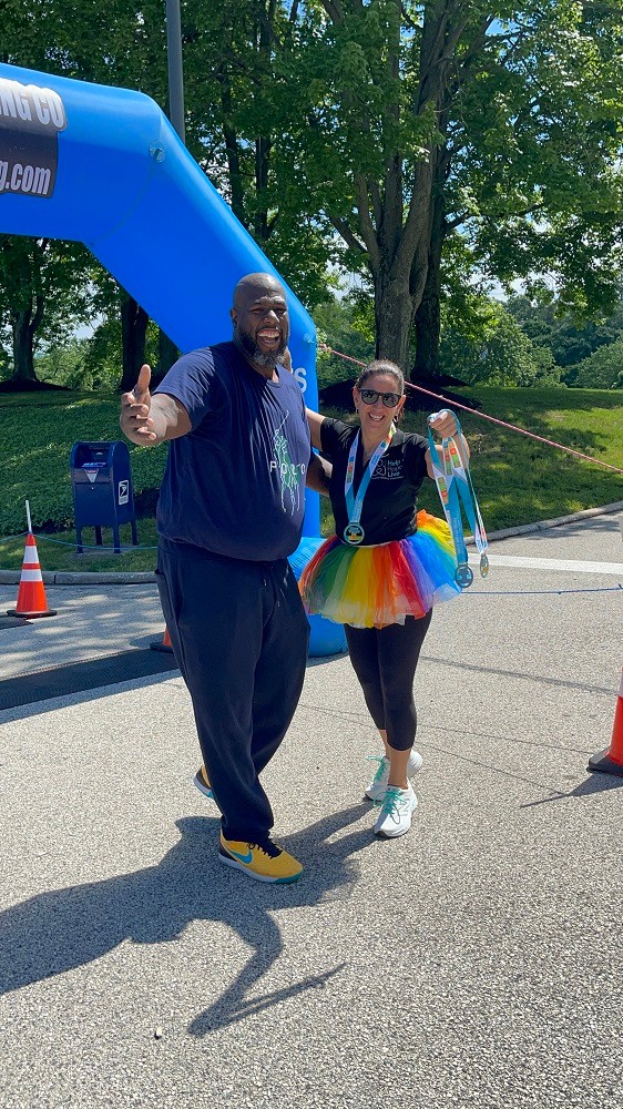 At Hope Travels 2024, Help Hope Live Executive Director Kelly L Green celebrates with Barrett Brooks. Kelly wears a Hope Travels medal and holds two more in her hand. She has light skin, dark hair, sunglasses, and a rainbow tutu. Barrett has brown skin and a short black and gray goatee and he stands about 2 feet taller than Kelly.