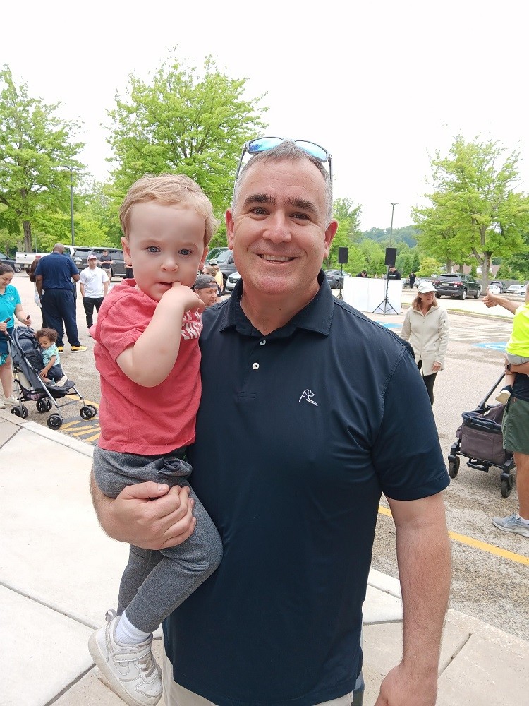 Help Hope Live board member Brian Messner holds his young son in his right arm outdoors at Hope Travels 2024. Brian has light skin, short gray hair, and a black polo shirt. His son has light skin, short red hair, and bright blue eyes.