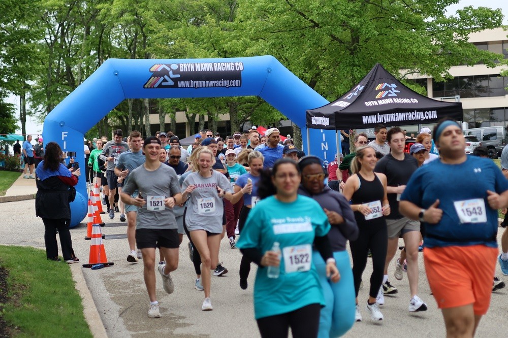 Dozens of runners take off from the Bryn Mawr Racing Co starting line at the Hope Travels 2024 5K event. They are a mix of ages, ethnicities, and genders wearing summer-appropriate athletic gear with runner numbers on their t-shirts.