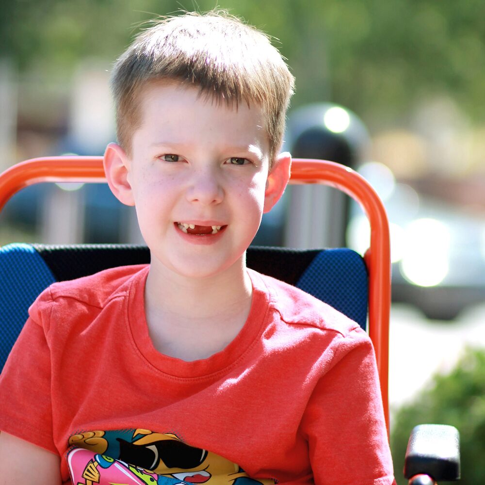 In a close-up outdoor portrait, Carson has light skin, short light brown hair, and green or blue eyes with his two front teeth missing. He wears a red t-shirt and is seated in a red, blue, and black power chair or mobility device.