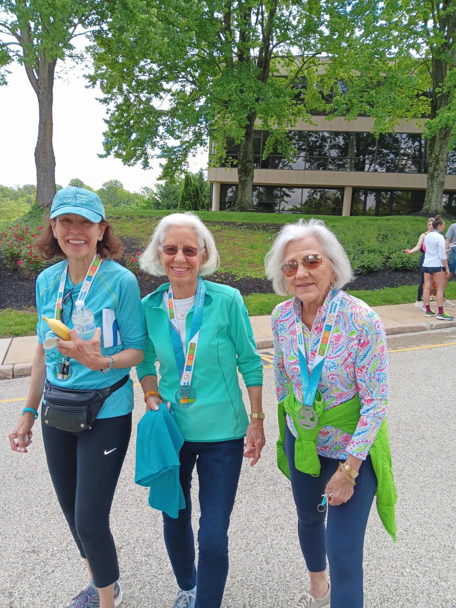 Three women pose for a photo during the Hope Travels 2024 event wearing athletic gear and their Hope Travels 5K medals around their necks. The first and third women are Help Hope Live legacy supporter Chris Kanter and co-founder Pat Kolff. Chris has light skin, shoulder-length brown hair, and a teal Help Hope Live cap. Pat has light skin, shoulder-length white hair, hoop earrings, and brown sunglasses.