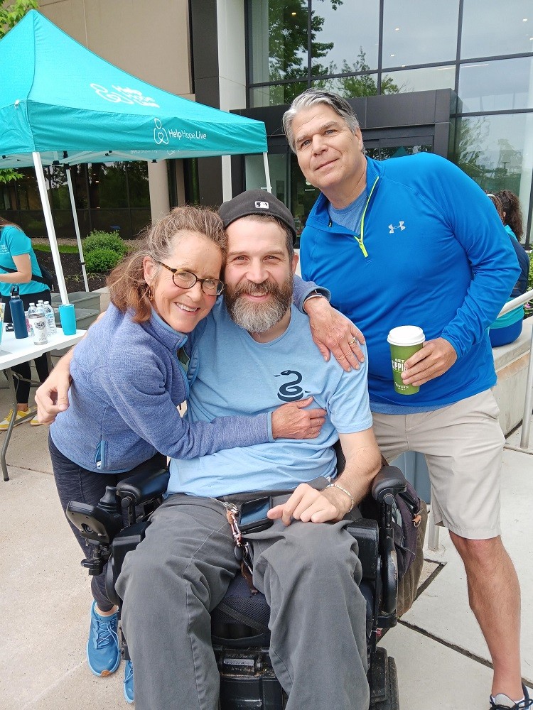 Former Help Hope Live Executive Director Lynne Samson with board member Jim Madden and client Jeff Harris. Lynne has light skin, tortoiseshell glasses, and gray-streaked brown hair as she stretches her arms around Jeff. Jeff has light skin, blue eyes, and a gray and brown beard and he is seated in his black power chair. Jim has light skin, gray hair, blue eyes, and athletic gear. They are outdoors at Hope Travels 2024.