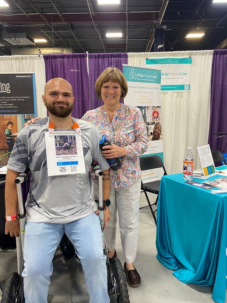 Beside the teal-themed Help Hope Live booth at Abilities Expo NY Metro 2024, Jack Perme stands in an assistive mobility device with treaded wheels beside Monica Perme. Jack has light skin, a shaved head, dark eyes, a neat short brown beard, and a big smile and he wears a laminated sign around his neck to advertise the Moving Forward Compassion Fund with Help Hope Live. Monica has light skin, a sandy bob with bangs, a flowery blouse, and khaki pants.