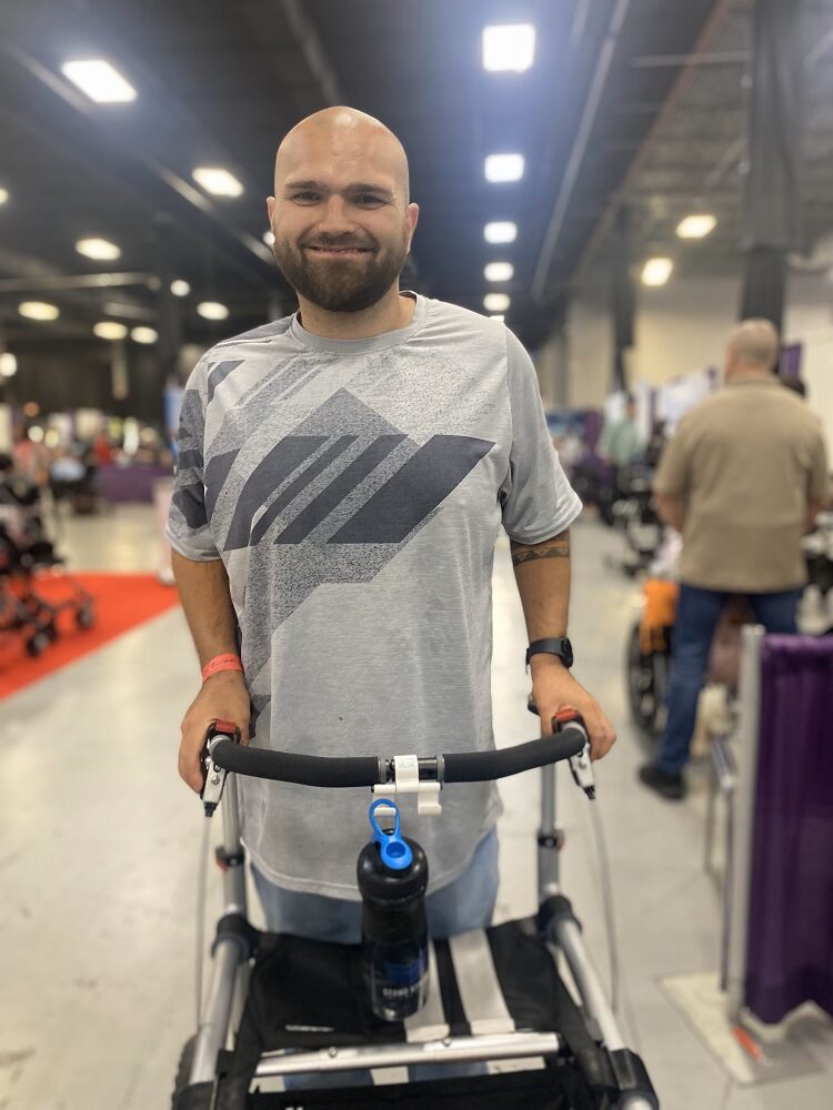 Standing with a black and silver mobility assistive device at Abilities Expo NY Metro 2024, Jack Perme has light skin, a shaved head, dark eyes, a neat short brown beard, and a big smile.