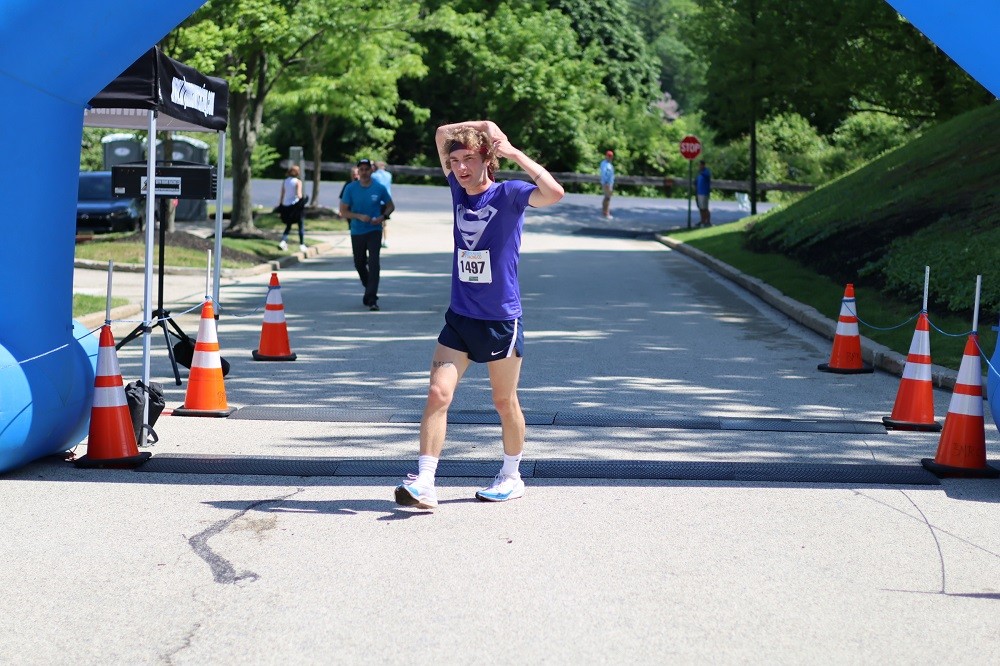 First place Hope Travels 5K 2024 finisher Jake Lawson stretches as he crosses the finish line outdoors. Jake has light skin, curly brown hair, a purple Superman logo t-shirt, and athletic shorts with sneakers.