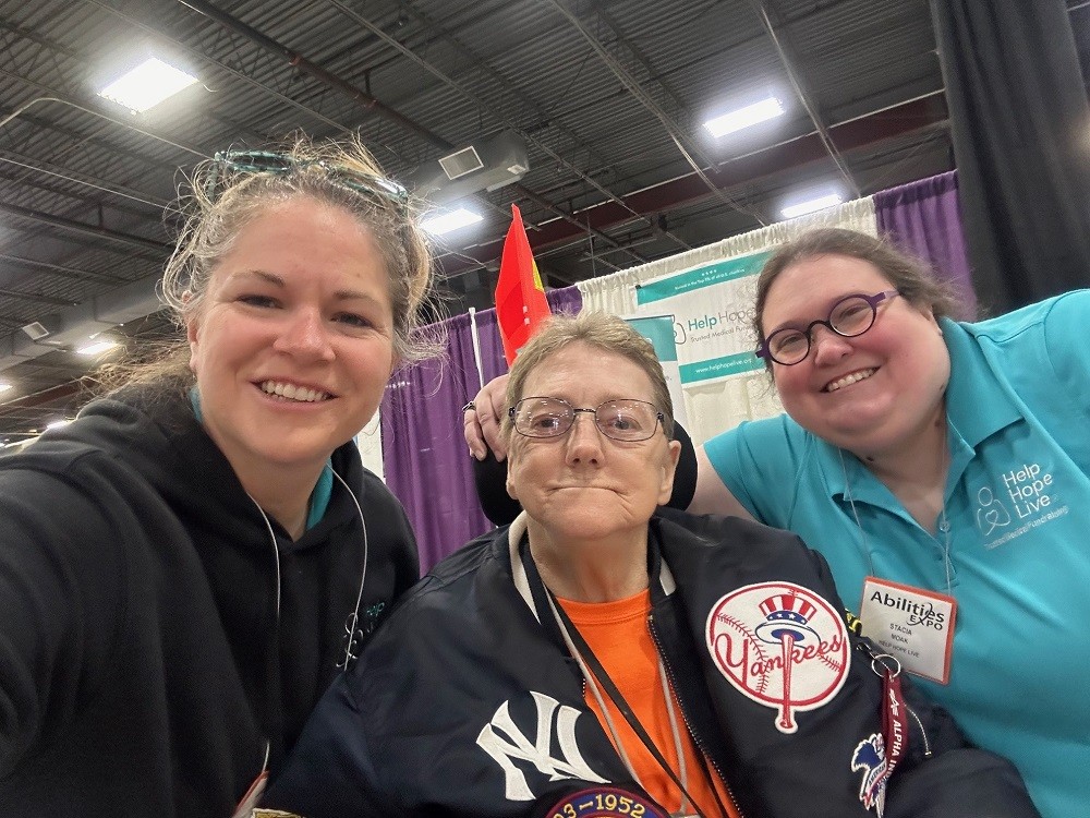 A selfie at Abilities Expo NY Metro 2024 with Sonny and Stacia from Help Hope Live and client Jane Koza. Stacia has light skin, round purple glasses, and curly brown hair and she wears a teal Help Hope Live polo shirt. Sonny has light skin, golden-brown straight hair, and a black Help Hope Live Nike fleece. Seated in a power chair, Jane has light skin, dark eyes, silver rimmed glasses, and short sandy hair, and she is decked out in NY Yankees sports team gear.