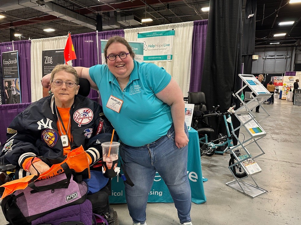 At Abilities Expo NY Metro 2024, Client Services Coordinator Stacia of Help Hope Live meets client Jane Koza. Standing, Stacia has light skin, round purple glasses, and curly brown hair and she wears jeans and a teal Help Hope Live polo shirt. Seated in a power chair, Jane has light skin, dark eyes, silver rimmed glasses, and short sandy hair, and she is decked out in NY Yankees sports team gear.