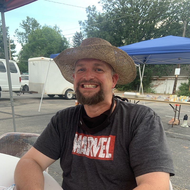 Kidney transplant recipient Josh Raymond is seated at an outdoor event or fundraiser. He has light skin, blue eyes, gray hair, a dark beard, and a big grin and he wears a straw cowboy-style sunhat and a MARVEL logo t-shirt.