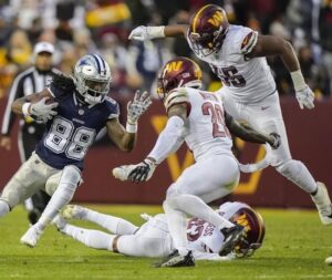 An action shot of KJ Henry and his teammates on the field of an NFL game. KJ plays for the Washington Commanders and he and his teammates are wearing Commanders uniforms in signature white, maroon, and gold on the bright green field.