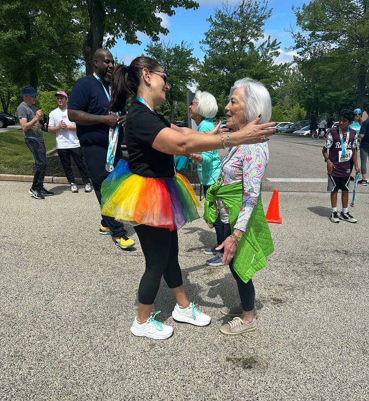 Current Help Hope Live Executive Director Kelly L Green reaches for co-founder Pat Kolff to pull her into a hug outdoors at Hope Travels 2024 with Barrett Brooks visible behind them. Kelly has light skin, dark hair in a ponytail, sunglasses, and a rainbow tutu. Pat has light skin, athletic clothing, and shoulder-length white hair. Barrett has brown skin and navy athletic gear and holds Hope Travels 5K medals to distribute.