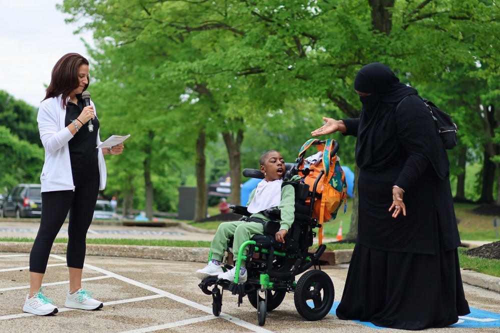 Outdoors at Hope Travels 2024, Help Hope Live Executive Director Kelly L Green speaks into a microphone in front of 8-year-old Khuwaylid Seeney and his mother. Khuwaylid has brown skin and is seated in his black wheelchair with lime green accents. His mother behind him wears a niqab in black with her eyes and hands visible. She has brown skin and is raising her arms in excitement as she addresses Khuwaylid. Kelly has light skin, brown hair past her shoulders, and a black Help Hope Live polo.