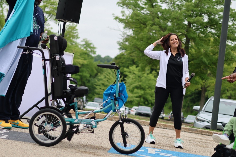 Outdoors at Hope Travels 2024, Help Hope Live Executive Director Kelly L Green grins as Barrett Brooks unveils a teal customized three-wheeled adaptive bike. Kelly has light skin, brown hair past her shoulders, and a black Help Hope Live polo and she holds a microphone in her left hand.