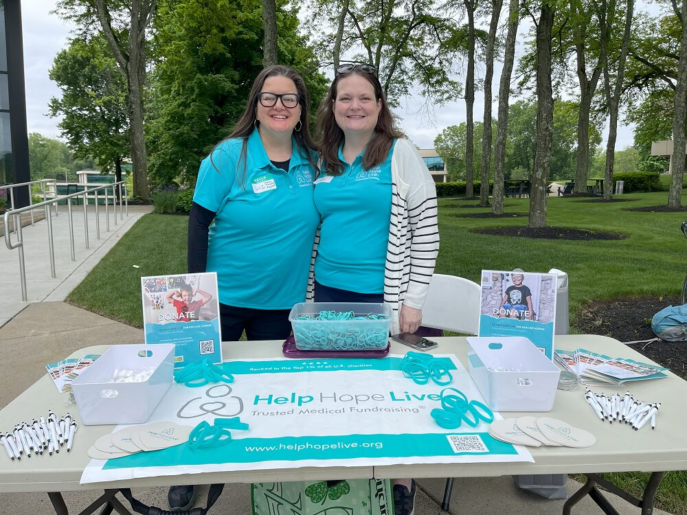 Help Hope Live client ambassadors Liz Casperite and Linda Jara wear matching Help Hope Live teal polos as they stand behind the Help Hope Live info table outdoors at Hope Travels 2024. Liz has light skin, black glasses, hoop earrings, and long gray-streaked brown hair. Linda has light skin, brown hair past her shoulders, and blue eyes. On the table is a Help Hope Live Trusted Medical Fundraising banner with teal bracelets, brochures, pens, signs, and more.