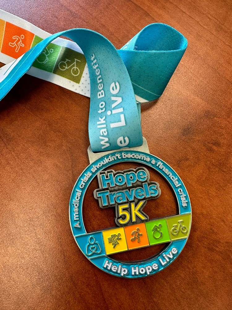 A close-up photo of a Hope Travels medal from Hope Travels 2024. The medals says Hope Travels 5k, A medical crisis should never become a financial crisis, Help Hope Live and features details in teal, white, yellow, orange, and green.