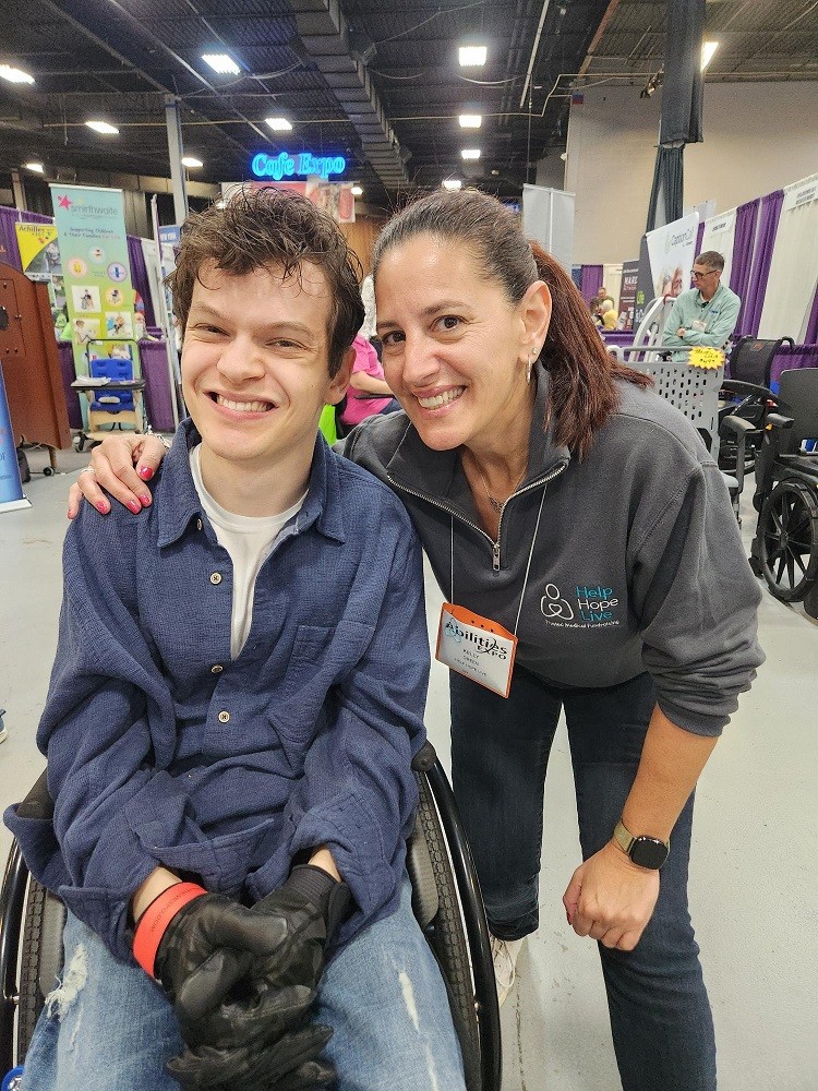 At Abilities Expo NY Metro 2024, Help Hope Live Executive Director Kelly L Green meets actor Micah Fowler. Kelly has light skin, brown eyes, dark hair in a ponytail, and a gray Help Hope Live branded sweatshirt with blue jeans. Micah is seated in his black manual wheelchair and has light skin, curly brown hair, brown eyes, a blue flannel, and black leather or faux-leather wheelchair gloves.
