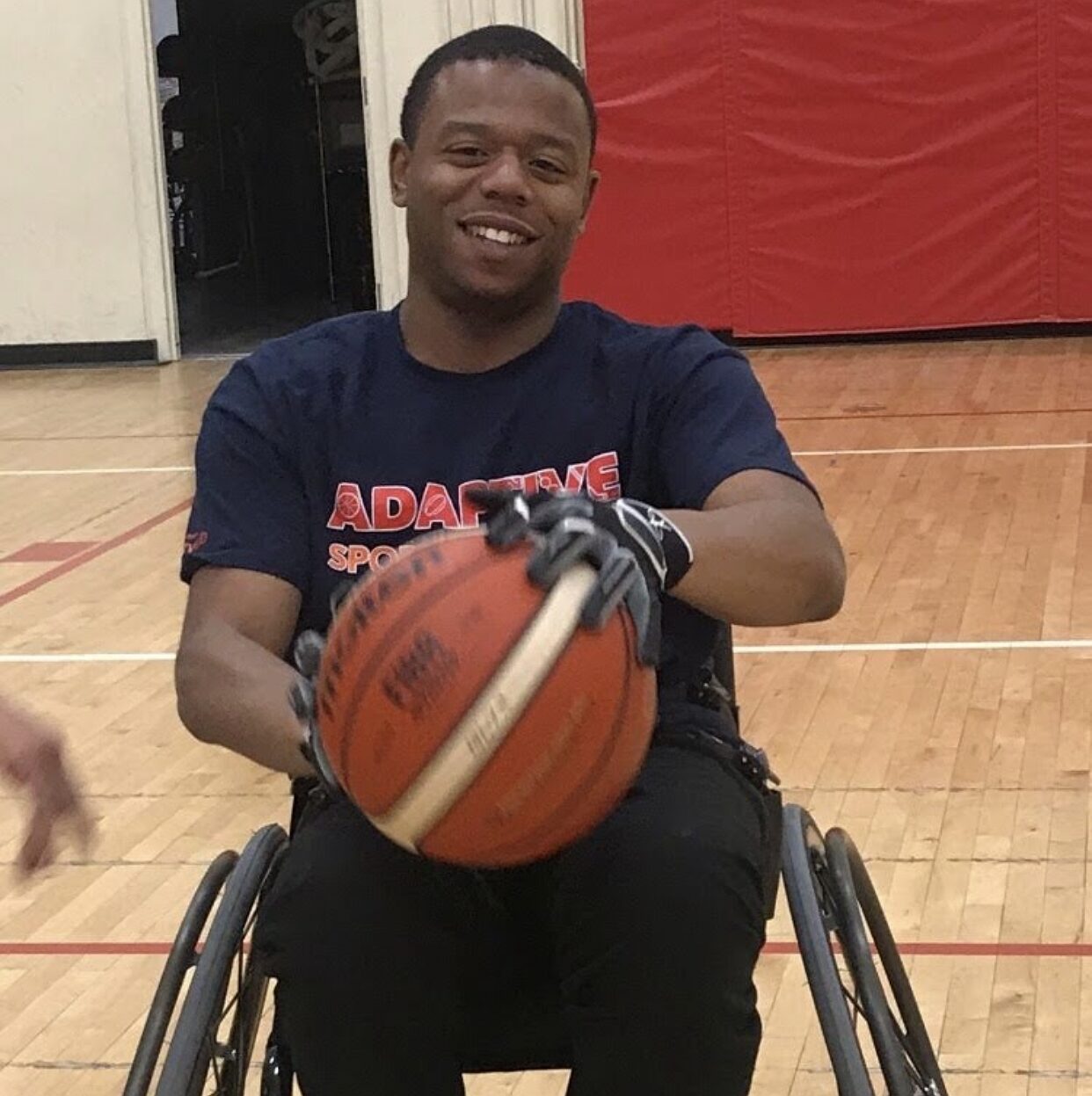 Michael Walthall is seated in his black wheelchair with a big smile on a basketball court as he holds a basketball in gloved hands. Michael has brown skin and short black hair.