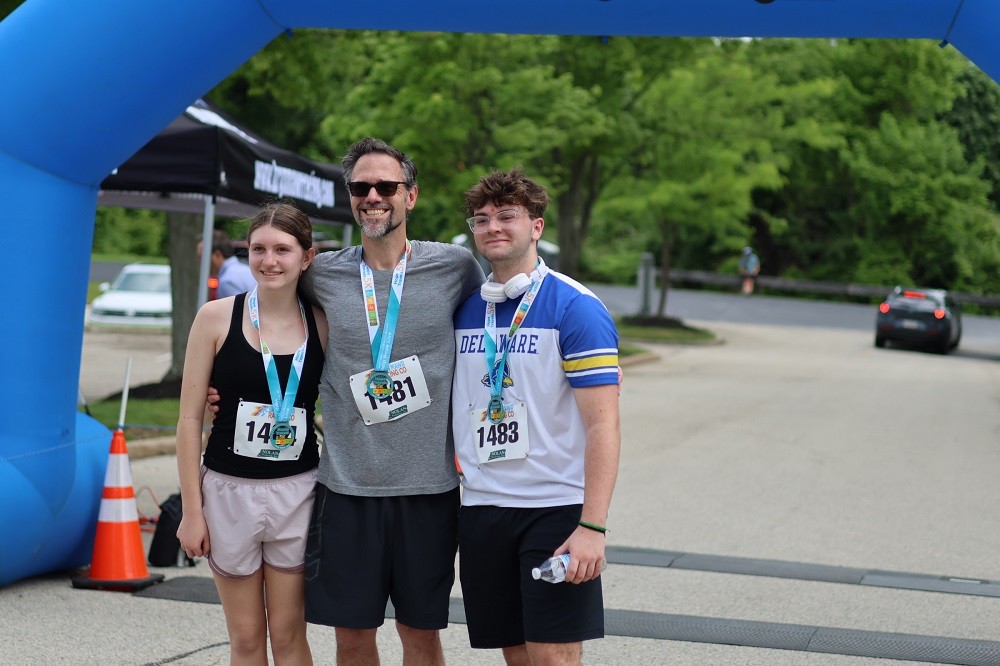 Help Hope Live board member Michael Criscuolo poses with two teens at the Hope Travels 2024 finish line outdoors. All three sport athletic gear appropriate for summer and Hope Travels medals with their runner numbers on their shirts. Michael has light skin, gray and black hair, and a gray and black goatee.