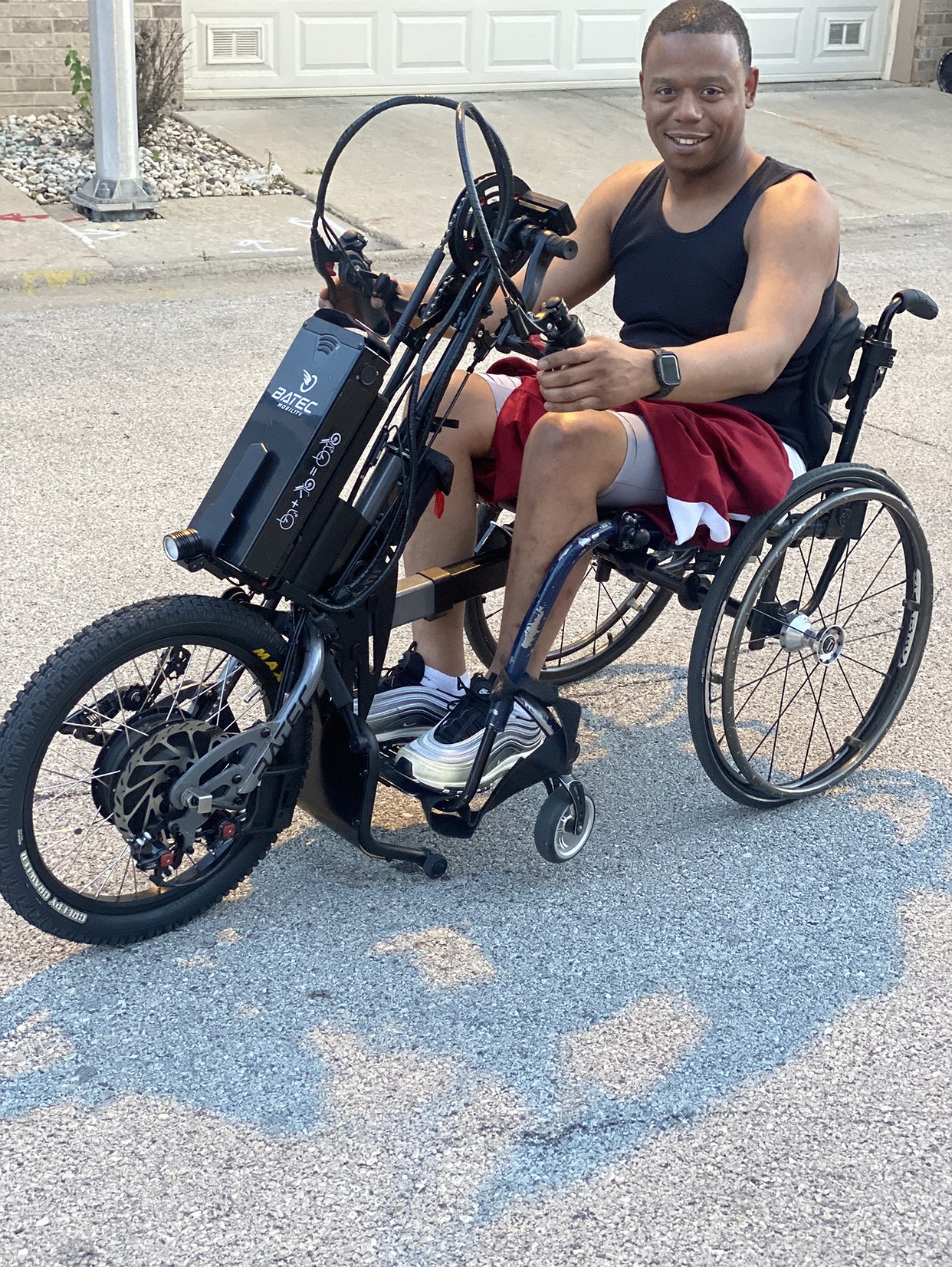 Michael Walthall is pictured outdoors in a street seated in his wheelchair with his feet and hands on a Batec adaptive mobility bike that protrudes from the front of his wheelchair with a large front wheel. Michael has brown skin, short black hair, and athletic warm weather clothing.