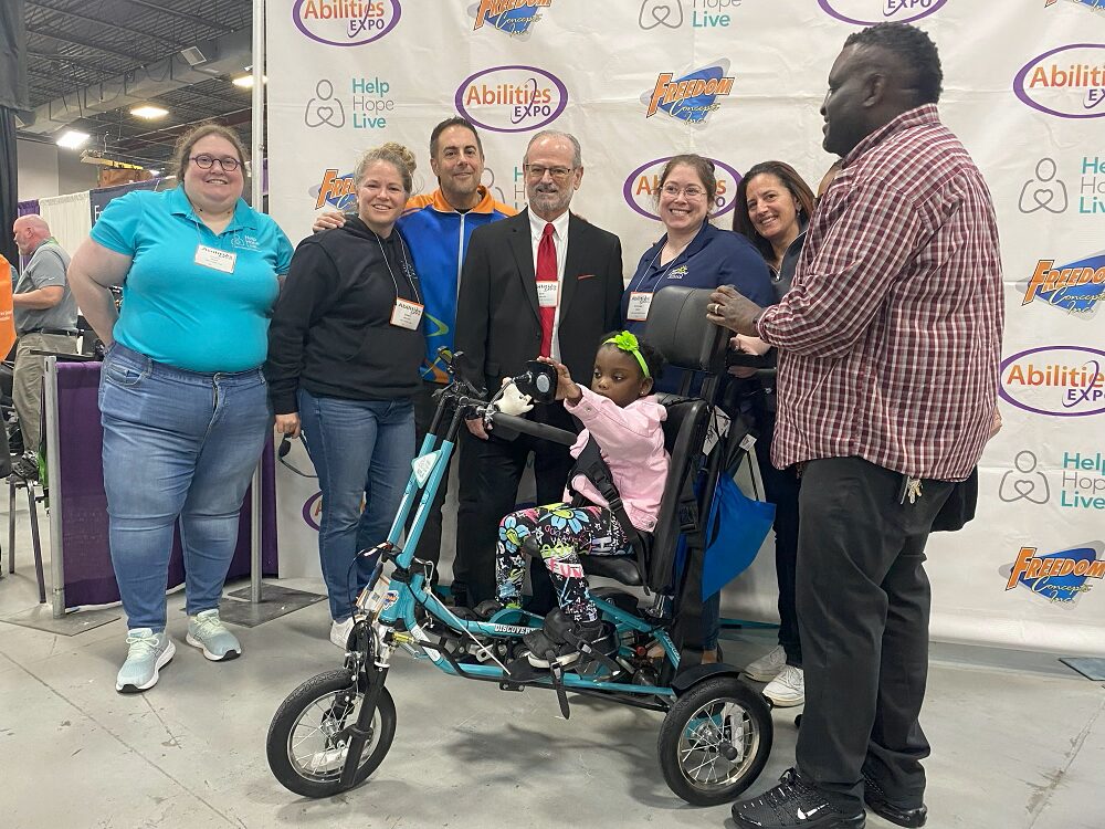 Seven people stand around 5-year-old Nyla Richmond on her new adaptive tricycle in front of a banner with logos for Help Hope Live, Freedom Concepts, and Abilities Expo. They are at Abilities Expo NY Metro 2024. The attendees include four women, three of whom work for Help Hope Live - Stacia, Sonny, and Kelly L Green - and three men, one wearing a suit with light skin and glasses, one wearing orange and blue Freedom Concepts themed clothing with light skin and short dark hair, and Nyla's father, who has brown skin and dark curly hair. Stacia has light skin, curly hair, purple circular glasses, jeans, and a Help Hope Live teal polo shirt. Sonny has light skin, a black Help Hope Live hoodie, jeans, and golden-brown hair pulled back. Kelly has light skin, a gray sweater, and straight red-brown hair. The fourth woman pictured has light skin, glasses, brown hair, and a navy polo. Seated on her teal and black new adaptive bike, Nyla has brown skin, dark eyes, short curly dark hair, a vivid laser-green headband with a flower, a pink collared top, and graffiti-style printed leggings plus stud earrings.