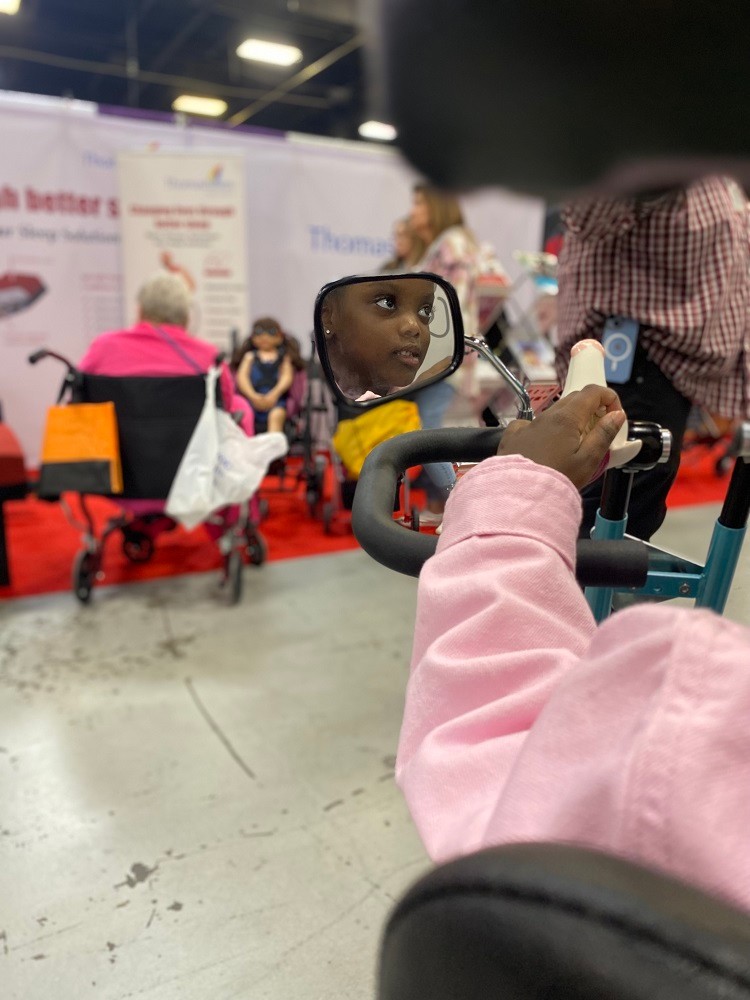 An artistic photograph taken over her shoulder shows the reflection of 5-year-old Nyla Richmond in the side mirror of her new Freedom Concepts adaptive tricycle at Abilities Expo NY Metro 2042. Nyla has brown skin, dark eyes, and stud earrings visible in the small mirror.