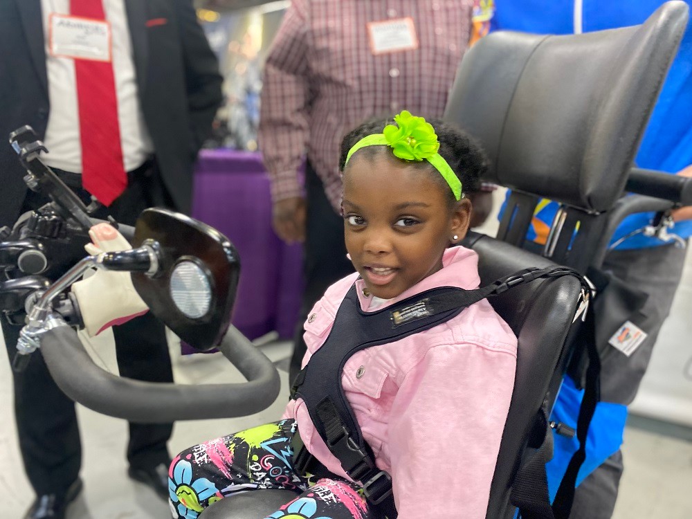 Seated on her new black adaptive tricycle from Freedom Concepts at Abilities Expo NY Metro 2024, 5-year-old Nyla Richmond smiles. She has brown skin, dark eyes, short curly dark hair, a vivid laser-green headband with a flower, a pink collared top, and graffiti-style printed leggings plus stud earrings.