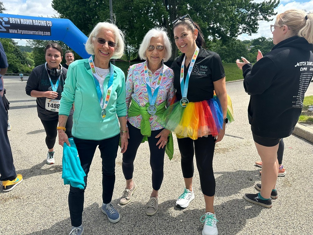 A photo of three women next to the Bryn Mawr Racing Co starting line at Hope Travels 2024. Two of them are significant Help Hope Live supporters: current Help Hope Live Executive Director Kelly L Green, who has light skin, dark hair pulled back, sneakers, a black Help Hope Live polo, and a rainbow tutu – and co-founder Pat Kolff, who has light skin, brown sunglasses, sneakers, and white shoulder-length hair. The three women wear Hope Travels 5K medals around their necks.