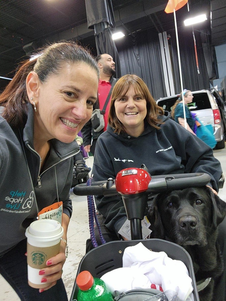 A selfie with Help Hope Live Executive Director Kelly L Green, client Amy Sherwood, and her service dog Dolly Pawton. Kelly has light skin, brown eyes, dark hair in a ponytail, and a gray Help Hope Live branded sweatshirt. She holds a Starbucks cup in her left hand. Amy Sherwood is seated on a mobility scooter and she has light skin, blue or hazel eyes, and shoulder-length straight red-brown hair with bangs. Dolly Pawton is a black labrador.