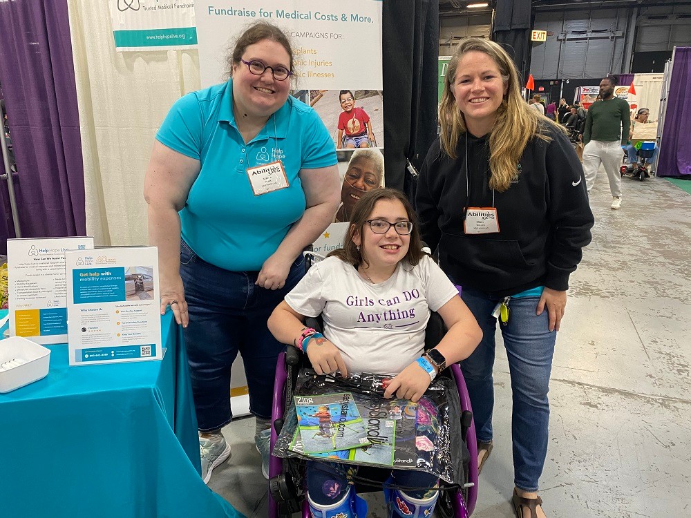 Stacia and Sonny from Help Hope Live with young client Laurel Jastrzembski at Abilities Expo New York Metro 2024. On the expo floor beside the teal-tableclothed Help Hope Live info booth, Sonny and Stacia are standing beside Laurel who is seated in her purple wheelchair. Stacia has light skin, round purple glasses, and curly brown hair and she wears jeans and a teal Help Hope Live polo shirt. Sonny has light skin, golden-brown straight hair, a black Help Hope Live Nike fleece, and jeans. Laurel has light skin, straight brown hair, black glasses, and a white t-shirt that reads Girl Can Do Anything in purple, and she wears blue leg braces decorated with stickers.