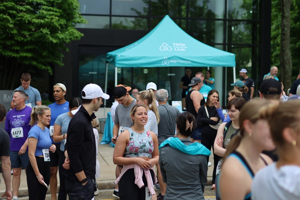 Hope Travels 2024 attendees mingle in a corporate parking lot with a Help Hope Live teal vendor tent visible. Several attendees are in athletic outfits with runner numbers on their shirts.
