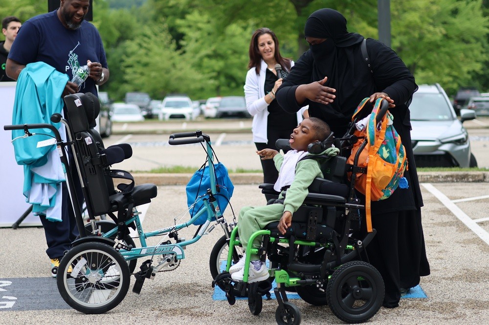 Outdoors at Hope Travels 2024, Help Hope Live Executive Director Kelly L Green and Barrett Brooks look on as 8-year-old Khuwaylid Seeney sees his customized teal adaptive bike for the first time. Khuwaylid has brown skin and is seated in his black wheelchair with lime green accents. His mother behind him wears a niqab in black with her eyes and hands visible. She has brown skin. Barrett has brown skin and a short black and gray goatee. Kelly has light skin, brown hair past her shoulders, and a black Help Hope Live polo.